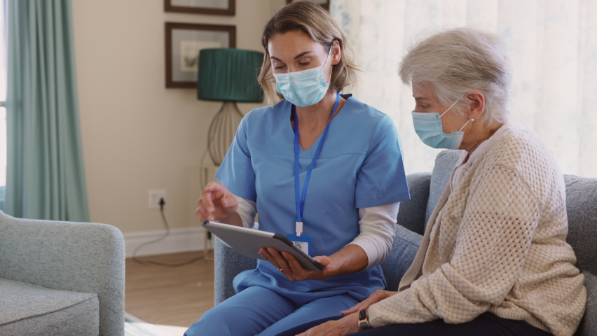 Young nurse and senior woman going through medical record on digital tablet during home visit and wearing face mask. Doctor during covid pandemic showing medical reports to old woman at nursing home.  | Shutterstock HD Video #1069200016