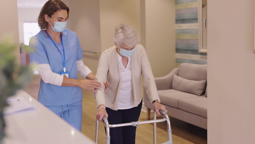 Smiling nurse with face mask helping senior woman to walk around the nursing home with walker. Young lovely nurse helping old woman with surgical mask for safety against covid-19 using a walking frame. Royalty-Free Stock Footage #1069200019