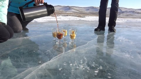 Blue ice of the lake, with cracks and bubbles. Hot mulled wine from a thermos is poured into glasses with chopped fruits. People's legs are visible. Background - snow-capped mountains. Baikal
Russia.