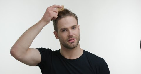 Portrait of Successful Young Man Combing Hair with Stylish Haircut. Smiling blonde Guy Looking Attractive at the Camera preparing for Workday wearing black T- Shirt. Daily Routine. Beauty Hair.