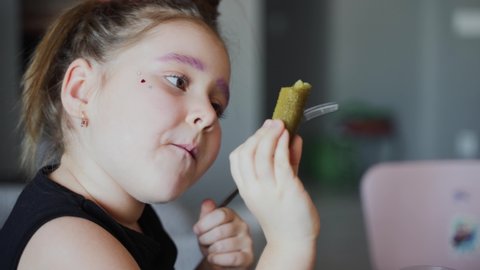 A authentic little girl is eating pickle at the table in the room with a fork and hand. Portrait of a kid eats with appetite and grimaces at home. Very fun video. Close up