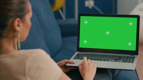 Woman sitting on couch in cozy living room working on laptop computer with mock up green screen chroma key display. Freelancer using isolated pc, browsing on internet for social media advertising