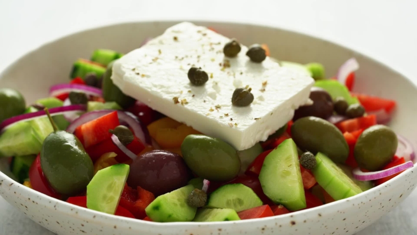 Sprinkling oregano seasoning to Greek village salad horiatiki with whole feta cheese and chopped vegetables, vegeterian mediterranean food, low calories keto diet, side view, close up, backlight