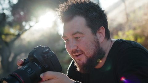 Professional photographer excited by creative photoshoot, concentrated on taking pictures in nature park on summer day at sunset. Slow motion handsome man looking in pro camera window and on screen