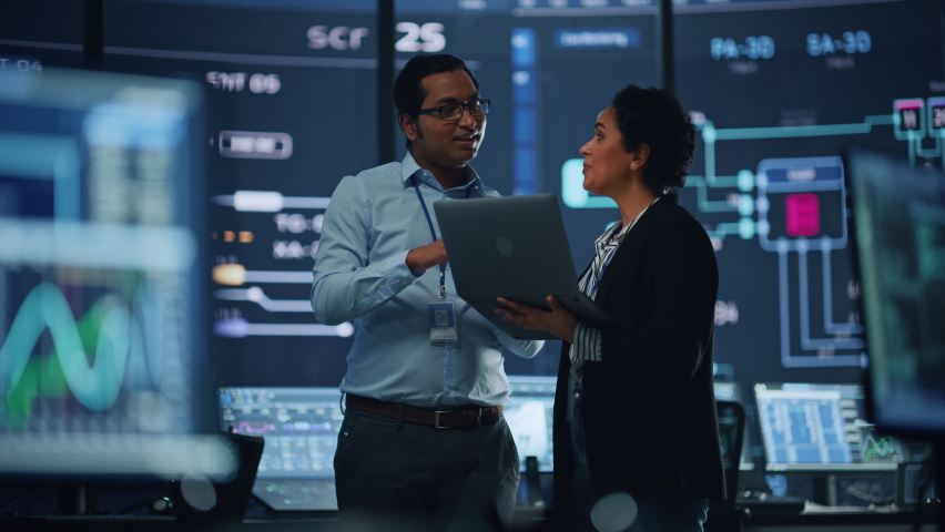 In the System Control Room Project Manager and IT Technical Engineer with Laptop Have Discussion, surrounded by Multiple Monitors with Graphics. Big Monitor Shows Interactive Server Blockchain Info. | Shutterstock HD Video #1069211413