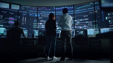In the System Control Room Project Manager and IT Technical Engineer Have Discussion, they're surrounded by Multiple Monitors with Graphics. Big Monitor Shows Interactive Server Blockchain Info.