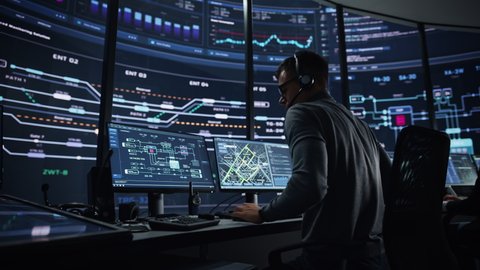 Professional IT Technical Support Specialist and Software Developer Working on Computer in Monitoring Control Room with Digital Screens. Employee Wears Headphones with Mic and Talking on a Call.
