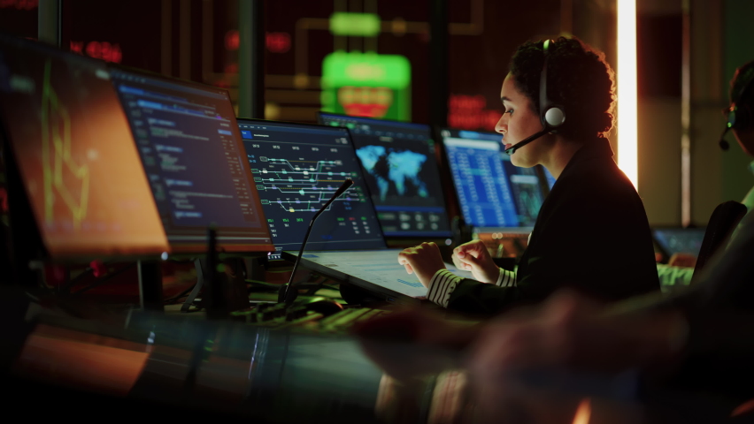 Multiethnic Female IT Technical Support Specialist Talking with a Client in Headphones and Working on Desktop Computer in Monitoring Control Room with Big Digital Screens with Server Blockchain Data. Royalty-Free Stock Footage #1069211491