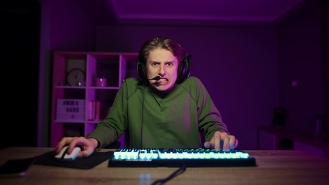 Evil gamer with a tense face plays video games at home on a computer in a room with purple lights and angrily looks at the camera. Emotional guy in a headset plays online games and streaming.