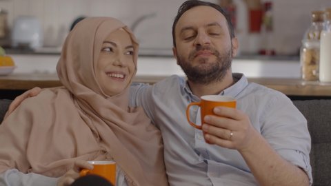 The Muslim woman with a hijab listens to her husband while drinking tea or coffee, and they chat and laugh. Concept of tea at homeClose up.