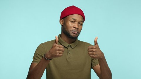Slow motion of cool Black man with beard, wearing red beanie, looking impressed with great work, showing thumbs up and pointing at camera, praise you, standing over blue background