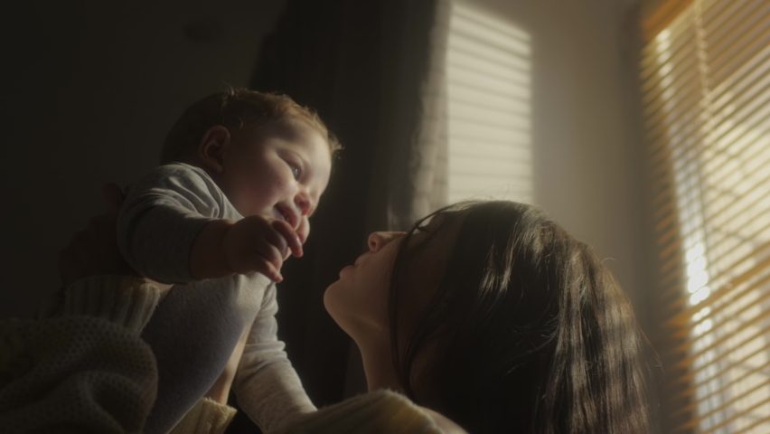 A child in the arms of a smiling girl. Millennial mother throws up in her son's arms at the window of the house. Blinds on the window. The child's face in the sun. Warm yellow tone. Family values. Royalty-Free Stock Footage #1069232353