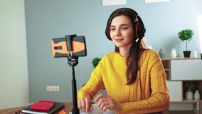 Happy young teacher in headphones communicates by video call using mobile phone. Woman in yellow sweater smiles and talks online while sitting table at home. Distance education and e learning concept