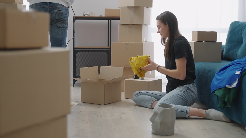 The newlyweds are unpacking things in their new apartment. The woman holds a yellow T-shirt in her hands and then puts it on the sofa with other things. High quality 4k footage