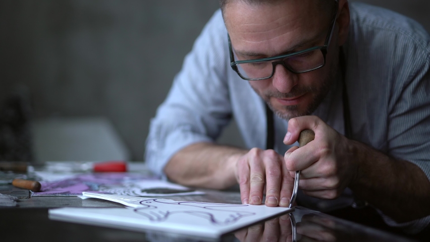 Linocut artist making manual carving process. Handmade work. Bearded middle aged man in apron making linocut using cutting instrument. 4k footage