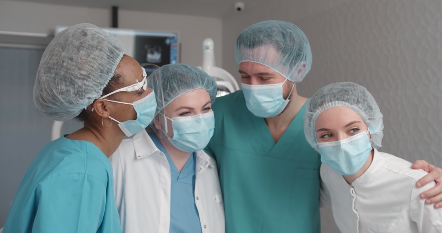 Portrait of doctors in uniform and safety mask hugging posing at camera in dental office. Diverse team of dentists embracing and smiling working in modern clinic | Shutterstock HD Video #1069242955