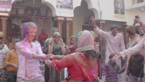 Two happy women a caucasian and traditional Indian performing spinning dance also known as 'Fugdi' are surrounded by people celebrating Holi festival with colours, Vrindavan, India (March 2020) 