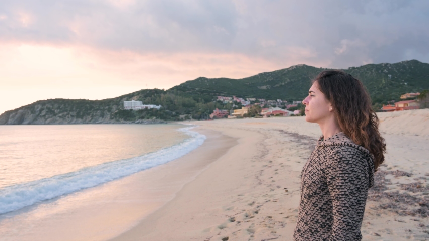 Side view young woman with jacket on the beach holding a camera and taking photo of the beautiful landscape during sunset.