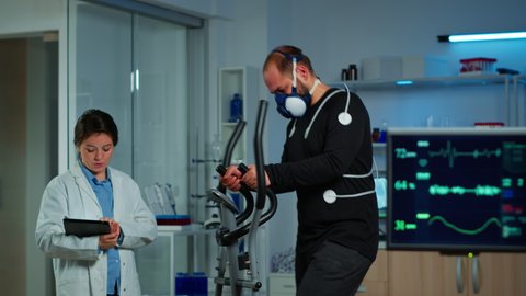 Patient making electrocardiogram during physical exercise in sports science lab, monitoring endurance and heart rate, computer showing EKG data on display. Man running with mask and medical electrodes