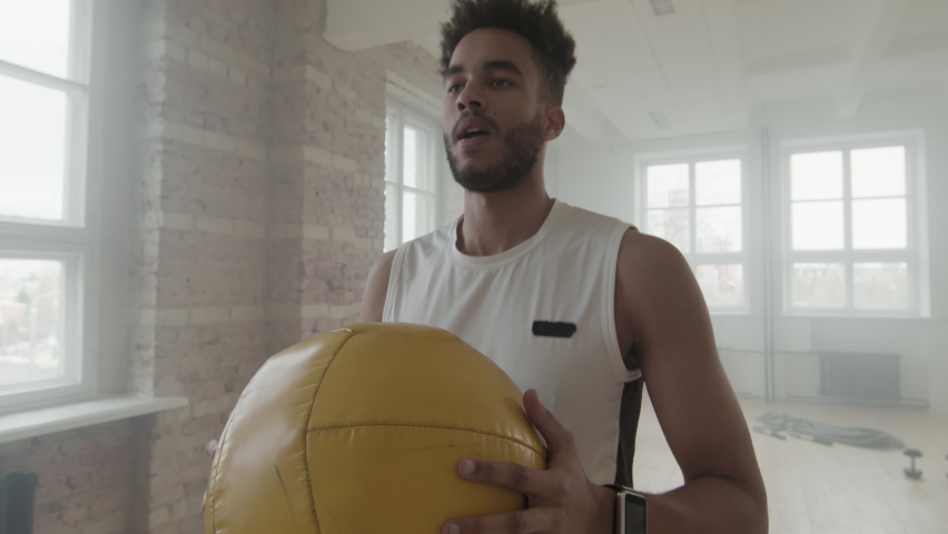 Low-angle medium shot with slowmo of concentrated african-american fit man doing squats with weight ball indoors
