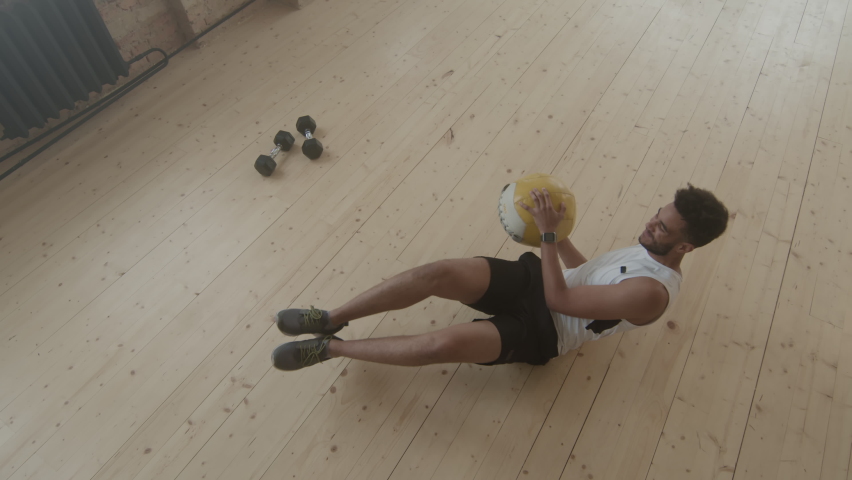 Top-view slowmo of fit afro-american man having abs workout with heavy weight ball lying on floor of smoky loft like gym studio