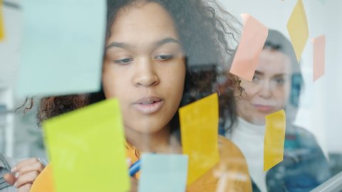 Close-up of Afro-American woman writing on sticky notes and discussing ideas with multi-ethnic team standing behind glass board in creative office