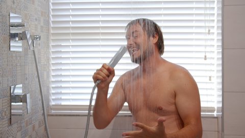 Young handsome man with dark hair takes a shower and emotionally sings like into microphone. Jets of water flow down the face and chest.