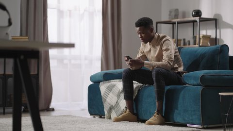 young black man is playing video game in smartphone, sitting at home alone, afro-american guy is communicating in social nets in mobile phone