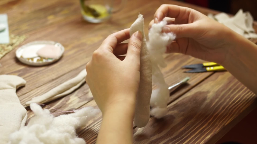 Woman fills detail for a rag doll with sintepon. Toys made of linen fabric. Mug on wooden table. Handmade diy work. | Shutterstock HD Video #1069291012
