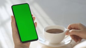Woman Hand Cup of Hot tea and Using Smartphone Watching Green Screen Top View. Smartphone with Green Mock-up Screen Business Concept.