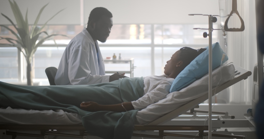African male doctor checking up female patient in hospital ward. Side view of black man practitioner talking to sick young woman lying in hospital bed and writing down in clipboard Royalty-Free Stock Footage #1069293526