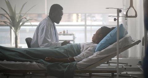 African male doctor checking up female patient in hospital ward. Side view of black man practitioner talking to sick young woman lying in hospital bed and writing down in clipboard