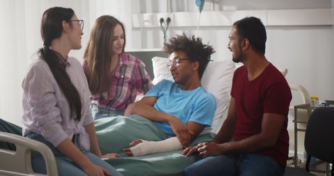 Diverse friends visit sick afro patient with broken arm in hospital. Portrait of young afro-american man with hand in plaster cast lying in hospital bed talking with diverse visitors friends