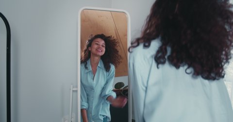 Rear view beautiful young smiling mixed race blogger woman touching curly hair, dancing enjoying her mirror reflection.