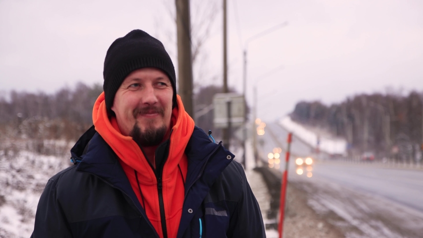 A serious young man standing on the side of the road begins to smile. A male in a warm jacket with an orange collar stands on the highway. In the background, trucks and cars are passing in defocus. | Shutterstock HD Video #1069296706
