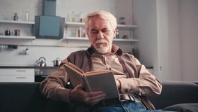 Senior man reading book while sitting on couch with kitchen on background