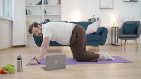 Humorous obese male doing Pilates at home, watching video on laptop, fitness
