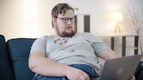 Disorganized overweight man in glasses typing on laptop, eating hot dog, hygiene