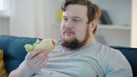 Untidy obese man eating hot dog at home, slovenly lifestyle, high cholesterol