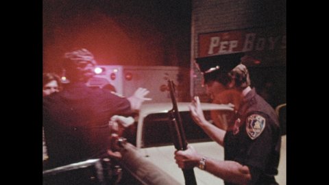 1970s: Crime scene. Police officer stashes rifle in cop car. A curly-haired detective walks up putting on a blazer and speaks with a cop. Paramedics gather around a dead man with a large bloody wound.