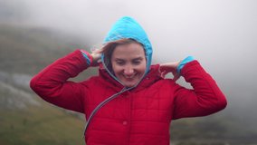 Girl hiker video portrait in the mountains, putting on hood. Cold weather with fog and strong wind in autumn.