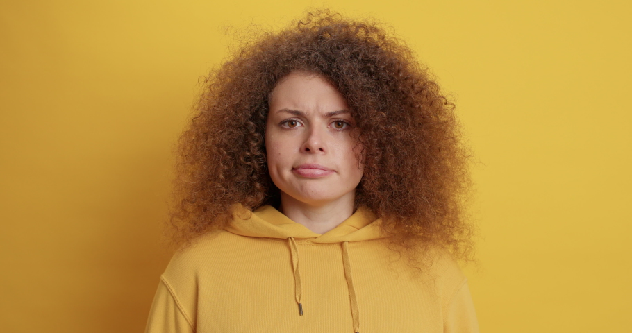 Doubtful curly haired serious woman purses lips has hesitant expression shakes head and says no dressed in sweatshirt isolated over yellow background. People hesitations and face expressions Royalty-Free Stock Footage #1069327876