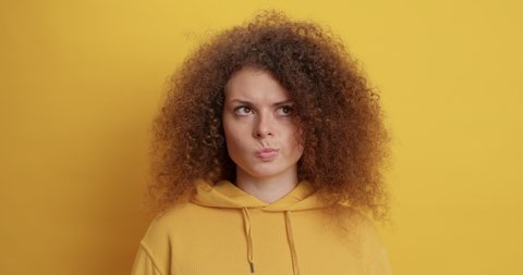 Doubtful curly haired serious woman purses lips has hesitant expression shakes head and says no dressed in sweatshirt isolated over yellow background. People hesitations and face expressions
