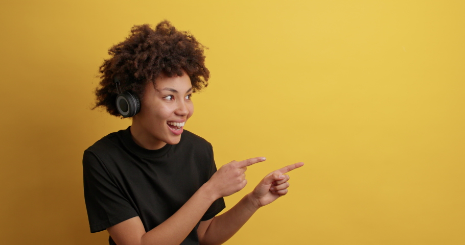 Cheerful African American woman indicates aside on copy space shows banner with product has surprised cheerful expression wears headphones on ears dressed in casual black t shirt. Wow how great | Shutterstock HD Video #1069328032