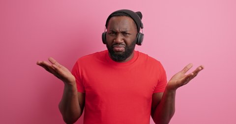 Displeased indignant bearded man spreads palms has no idea doesnt know what to do wears hat casual red t shirt isolated over pink background. Dissatisfied African American guy feels doubtful