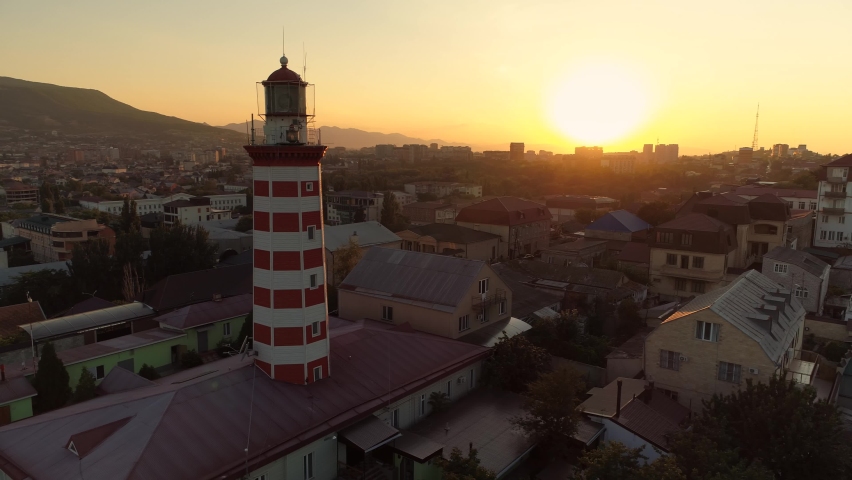 Makhachkala Petrovsky operating lighthouse Dagestan capital. Beautiful landmark at orange cinematic sunset. Sun rays shine through tower. Downtown scenic cityscape. Horizon. Aerial sideways flight. 4k