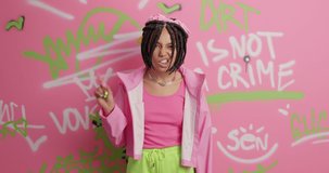 Youth subculture and urban lifestyle concept. Cool funky female teenager with dreadlocks raises index finger has self confident expression dressed in stylish clothes poses against graffiti wall