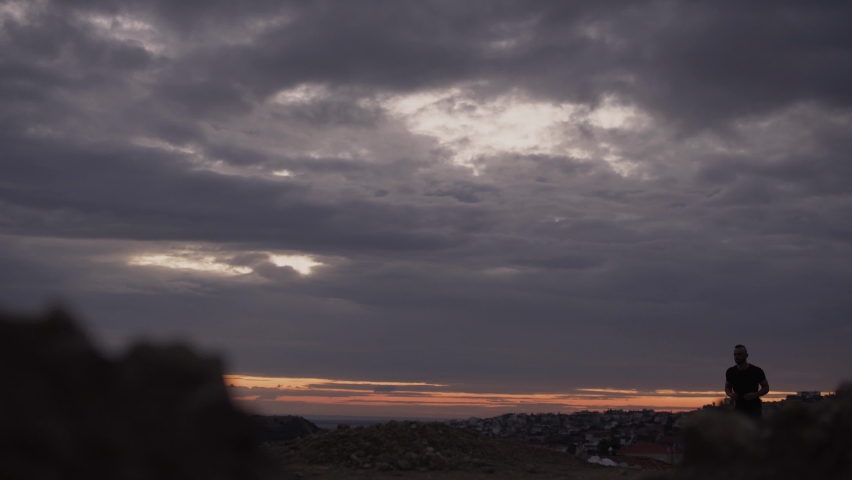Young athletic man jogging at sunset against city skyline and cloudy sky before dusk