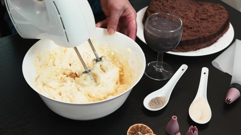 Woman is making chocolate cream, adding hot chocolate to bowl with butter. Female baker is whipping butter and hot cocoa chocolate using electric hand mixer. Process of making cake. Culinary concept