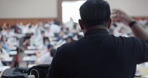 The male Asian teacher in black clothes teaches many Asian high school students in the auditorium using a laptop connected to a projector on a stage.